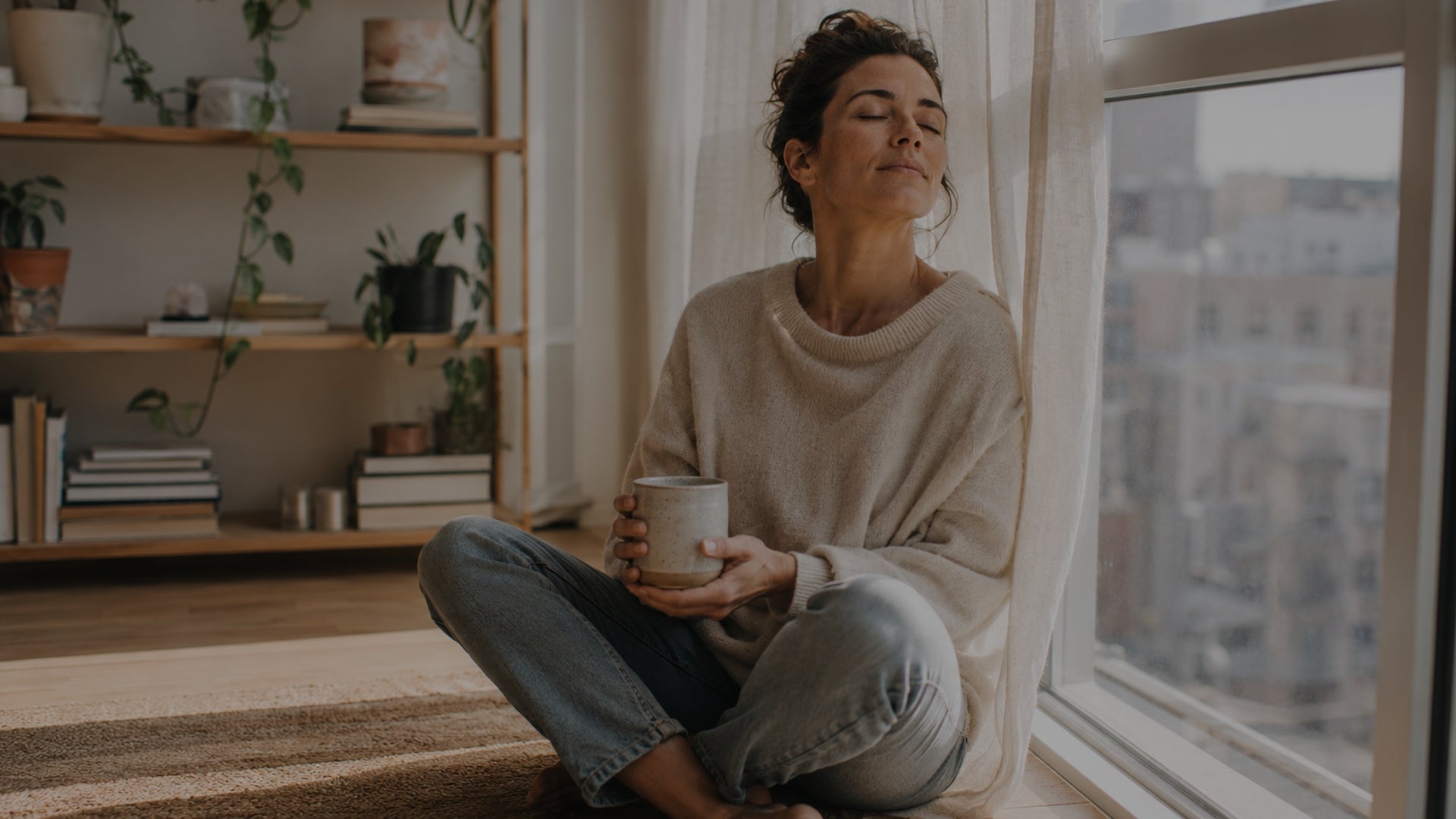 Woman sitting by a window holding a mug in a cozy room with a bookshelf.
