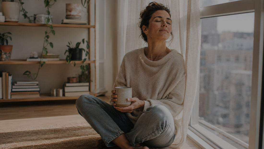 Woman sitting by a window holding a mug in a cozy room with a bookshelf.