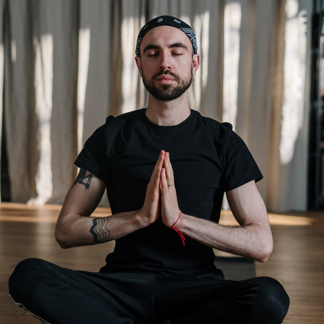 Man in black clothing meditating with hands pressed together in a room with curtains.